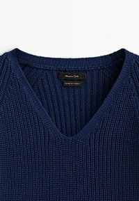 Marineblau gestrickter Pullover mit V-Ausschnitt, der ein strukturiertes Muster aufweist. Das Etikett trägt die Aufschrift „Massimo Dutti“, was darauf hinweist, dass er in der Türkei hergestellt wurde.