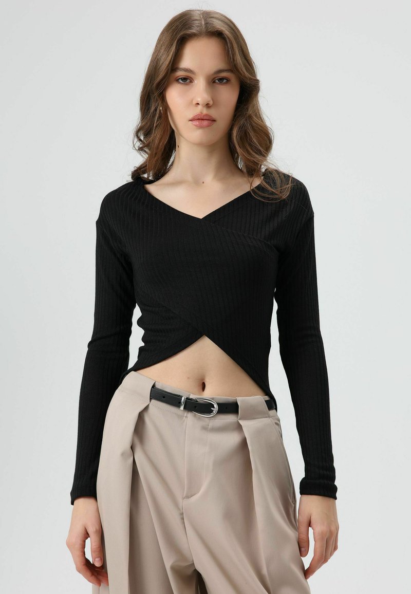 Zwarte geribbelde top met lange mouwen en een V-hals met een omwikkeld ontwerp, gecombineerd met lichtbeige broek met hoge taille, vastgemaakt met een zwarte riem.