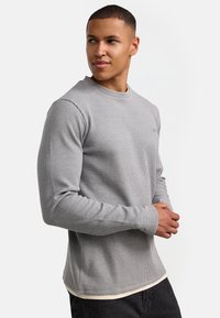 Suéter gris de manga larga con un patrón acanalado texturizado, cuello redondo y un ajuste relajado. Combinado con una camisa más clara debajo.