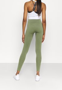 Leggings de talle alto en color verde oliva, fabricados con un tejido elástico, con una textura acanalada y combinados con zapatillas deportivas blancas.