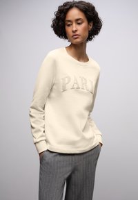 Cremesweatshirt mit dem Schriftzug „PARIS“ aus Plüschmaterial, Rundhalsausschnitt und gerippten Bündchen, kombiniert mit grauen Nadelstreifenhosen.