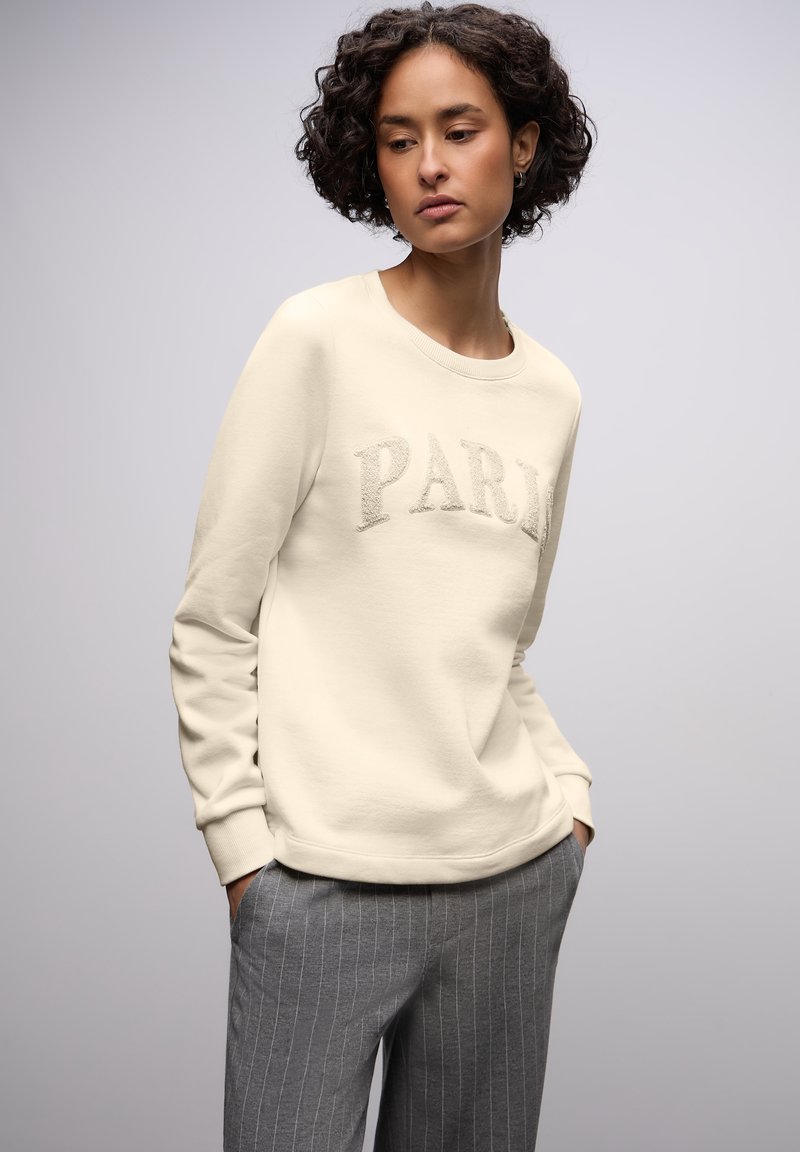 Cremesweatshirt mit dem Schriftzug „PARIS“ aus Plüschmaterial, Rundhalsausschnitt und gerippten Bündchen, kombiniert mit grauen Nadelstreifenhosen.