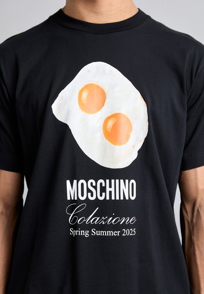 Black t-shirt featuring a white fried egg graphic with two orange yolks. The design includes "MOSCHINO Colazione Spring Summer 2025" in white text.
