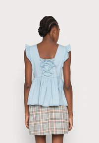 Top em chambray azul claro com mangas com babados, corpete justo e estilo peplum. As costas apresentam um detalhe de cordas. Combinado com uma saia xadrez.