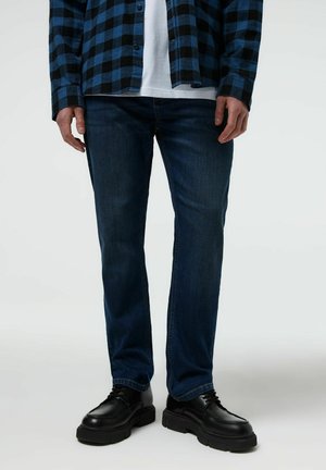 Mężczyzna ubrany w ciemnoniebieskie dżinsy, czarne masywne buty sznurowane, białą koszulę oraz niebiesko-czarną flanelową koszulę w kratę.