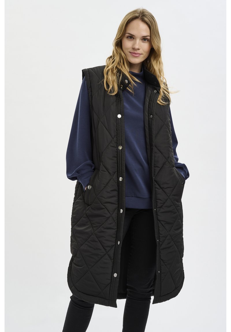 Veste sans manches matelassée noire au design long, avec des boutons-pression et des poches latérales, portée sur un pull bleu et un pantalon noir.