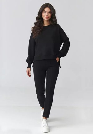 Kobieta o długich kręconych włosach, ubrana w czarną bluzkę z długim rękawem, czarne spodnie i białe trampki, idąca na tle jasnego, jednolitego tła.