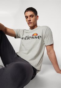 Camisa de algodón ligera de cuello redondo en gris claro con un audaz logotipo en negro de "ellesse" y un diseño de acento en rojo/naranja. Tejido suave con un corte relajado, combinada con pantalones negros.