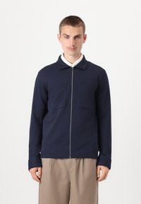 Navy zip-up jacket made of soft fabric with a collar, two chest pockets, and long sleeves, paired with beige trousers.