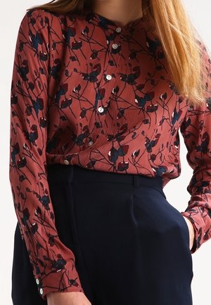 Femme portant un chemisier boutonné à fleurs rouge rouille rentré dans un pantalon noir taille haute, main dans la poche, avec des cheveux longs et raides visibles.