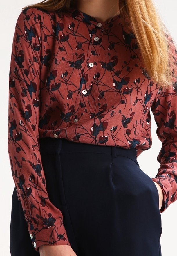 Femme portant un chemisier boutonné à fleurs rouge rouille rentré dans un pantalon noir taille haute, main dans la poche, avec des cheveux longs et raides visibles.