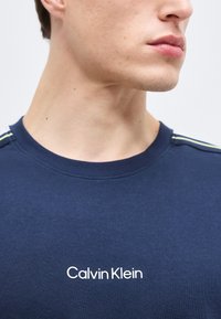 Κοντινό πλάνο ενός ατόμου που φοράει ένα μπλε ναυτικό t-shirt Calvin Klein με το λογότυπο στο κέντρο του στήθους και νέον ρίγες στους ώμους.