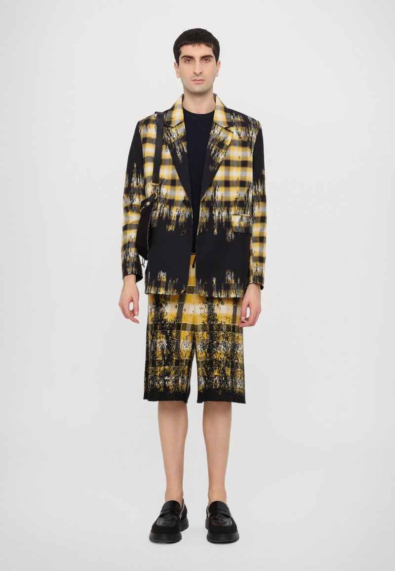 Black and yellow checkered blazer with a matching shorts set; features brushstroke patterns, a single button, and black shoes.