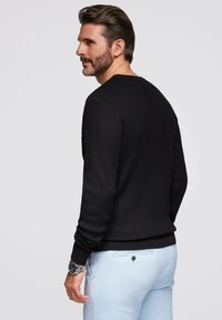 Maglione nero a coste con colletto rotondo e maniche lunghe, abbinato a pantaloni azzurri chiari. La texture è morbida e aderente. Orologio argento al polso.