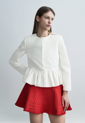 Kobieta o prostych, brązowych włosach, ubrana w biały, długi rękaw peplum i czerwoną plisowaną spódnicę na jednolitym jasnoszarym tle.