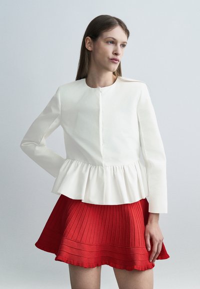 Femme aux cheveux bruns raides portant un haut péplum blanc à manches longues et une jupe plissée rouge sur un fond gris clair uni.
