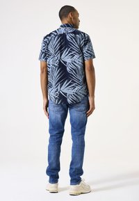 Camisa de manga corta de color marino con patrón de hojas de palma en azul claro; combinada con jeans azules claros y zapatillas crema; corte relajado, diseño casual.