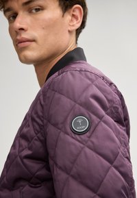 Gepufferde bomberjack in diep paars, met een zwarte ribgebreide kraag en een ronde logopatch op de linker mouw. Gladde textuur.