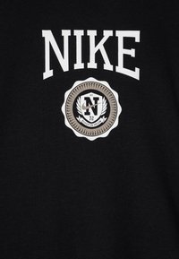 Melna jakas ar lielu baltu "NIKE" logo un apaļu emblēmu ar "N" un dekoratīviem elementiem zem tā. Mīksta, gluda tekstūra.