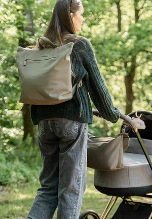 Moteris, apsivilkusi žalią megztinį ir nešanti kuprinę, stumia pilką vežimėlį su pritvirtinta krepšiu žaliai miško takeliu.