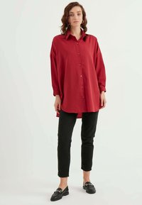 Rotes, langärmliges Hemd mit Knopfleiste und Kragen, mit einem lässigen Schnitt und seitlichen Schlitzen, kombiniert mit schwarzen Jeanshosen und schwarzen Slippern.