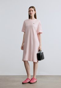 Ροζ βαμβακερό φόρεμα τύπου T-shirt με κοντά μανίκια, με ένα μικρό πράσινο λογότυπο. Συνδυάζεται με ροζ αθλητικά παπούτσια και κρατάει μια μαύρη τσάντα.