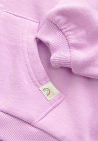 Zbliżenie na rękaw jasnofioletowej bluzy z prążkowanym mankietem i kieszenią kangura z małą beżową metką.