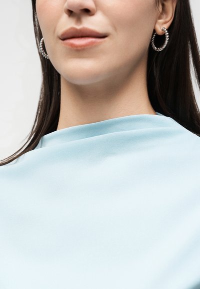 Γυναίκα που φοράει ασημένια στριφτά σκουλαρίκια κρίκους και ανοιχτό μπλε τοπ με υψηλό λαιμό, με ίσια σκούρα μαλλιά μερικώς ορατά.