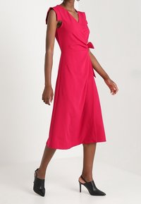 Robe portefeuille fuchsia avec manches courtes, ceinture à la taille et jupe fluide. Portée avec des escarpins noirs à bride arrière. Tissu lisse, longueur genou.