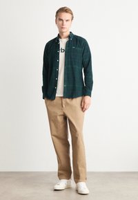 Grüne karierte Button-Up-Bluse über einem weißen T-Shirt, kombiniert mit beige Hosen und weißen Sneakers. Die Bluse hat eine Tasche.