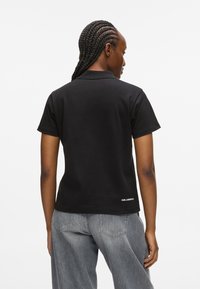 Fekete, rövid ujjú pólóing sima textúrával, klasszikus gallérral és egy kicsi fehér logóval az alján. Szürke farmernadrág látható.