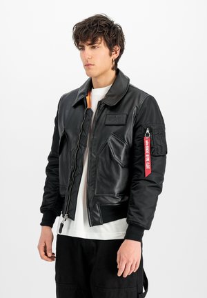 Jonge man met donker krullend haar die een zwart leren bomberjack draagt met een rode "Remove Before Flight" label op de mouw tegen een witte achtergrond.