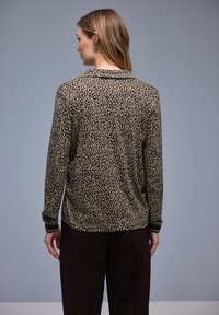 Chemise à manches longues avec un motif léopard beige et noir, dotée d'un col et de poignets côtelés. Portée avec un pantalon noir.