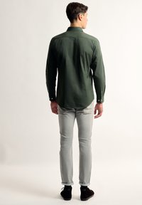 Camisa verde de manga larga con un corte recto, puños de botón y dobladillo redondeado, combinada con unos jeans gris claro y zapatos negros.