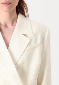 Blazer crema con un patrón floral texturizado, solapa de muesca, bolsillo en el pecho y silueta estructurada. La tela parece suave con un brillo sutil.