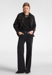 Giacca di denim nera con bottoni dorati, maniche lunghe, abbinata a una maglietta nera e jeans neri a gamba larga. Accento notevole: scarpe marroni a punta.