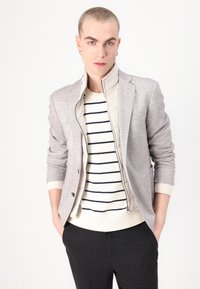 Blazer gris clair avec revers cranté, superposé à un pull crème à rayures. Pantalon sombre, texture de tissu lisse et fermeture éclair.