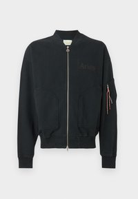 Chaqueta bomber negra con cremallera frontal, dos grandes bolsillos delanteros, logo de Aries en el pecho y bolsillo con cremallera y tiradores naranjas en la manga.