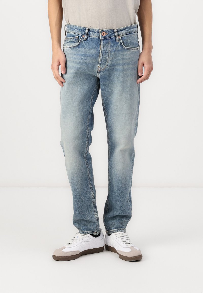 Jean en denim bleu clair avec une coupe droite, un délavage usé, cinq poches et une fermeture à bouton. Associé à des baskets blanches avec des accents gris.