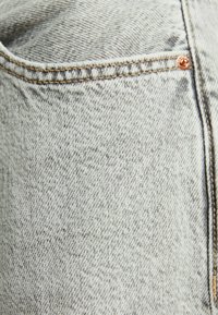 Gros plan d’un tissu denim gris clair avec des coutures détaillées et un rivet en cuivre sur l’angle d’une poche de jean.