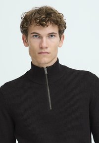 Pullover nero a maglia con colletto alto e dettaglio con zip corta al collo. Il tessuto presenta un motivo a coste testurizzato.