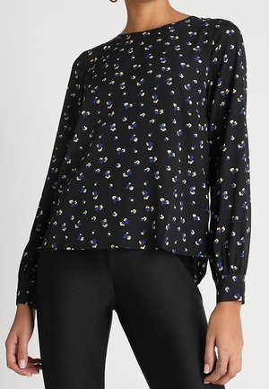 Vrouw draagt een zwarte blouse met lange mouwen met kleine blauwe en witte bloemenprints, gecombineerd met zwarte aansluitende broek.