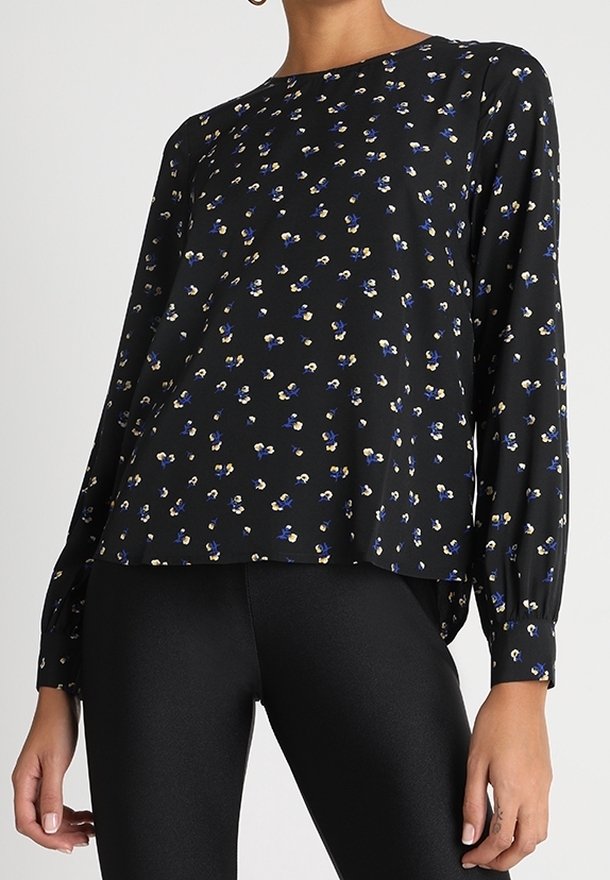 Femme portant un chemisier noir à manches longues avec de petits imprimés floraux bleus et blancs, associé à un pantalon noir ajusté.