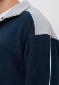 Dettaglio ravvicinato di una giacca in pile blu navy e grigio chiaro con dettagli in bianco sullo spallaccio e sulla parte superiore del braccio.