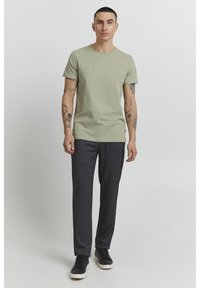 Grønn bomull T-skjorte med rund hals og preget logo, sammen med mørkegrå skreddersydde bukser og svarte sneakers med hvite såler.