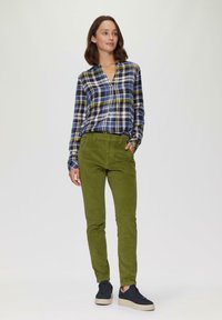 Geruite blouse met knopen in blauw, groen en wit; gecombineerd met groene corduroy broek en zwarte sneakers met een witte zool; model in staande pose.