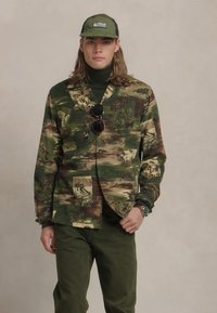 Jachetă de camuflaj cu modele verzi, maronii și bej; asociată cu un pulover negru cu guler înalt și pantaloni verzi. Accesoriile includ ochelari de soare și o șapcă.