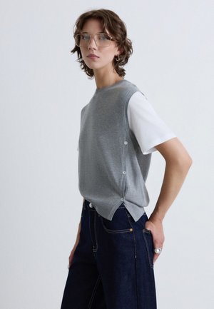 Jeune personne aux cheveux courts et grandes lunettes portant un pull sans manches gris sur un t-shirt blanc et un jean bleu foncé sur fond uni.