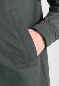 Main dans la poche latérale d'une veste gris foncé avec des poignets côtelés sur les manches.