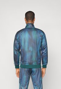 Chaqueta deportiva con un cuello alto, compuesta de un tejido ligero y texturizado. Presenta un patrón abstracto en tonos azul-verde y puños acanalados.